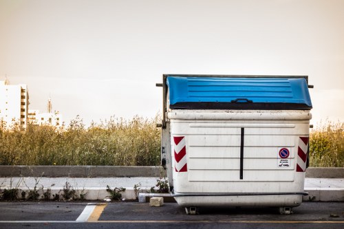 A large outdoor wheeled rubbish skip with a blue lid, positioned on a paved surface adjacent to a curb in a suburban or industrial area in Mortlake. The container is made of metal with visible horizontal ridges and has red and white reflective chevron markings on its sides for visibility. The skip features a small no-parking sign affixed to its front panel. Surrounding the skip are patches of grass and weeds along the roadside, with a background of tall, dense shrubbery and a partly cloudy sky, suggesting late afternoon or early evening lighting. The scene depicts typical rubbish collection infrastructure, relevant to professional waste management and rubbish removal services provided by Office Clearance Mortlake, targeting local businesses or residents in the Mortlake area associated with the postcode postcode. The environment indicates an active waste disposal or clearance site near residential or commercial premises in south-west London.
