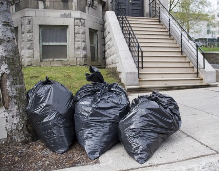 Three large black plastic rubbish bags filled with waste are positioned on the pavement near the base of a tree, in front of a building with a stone facade and a staircase leading to a raised entrance. The bags appear tightly knotted at the top, and their plastic surface is slightly crinkled and reflective. The bags are situated on a concrete sidewalk next to a grassy area with a tree trunk visible on the left. In the background, part of a staircase with white steps and black metal handrails ascends to a door or entrance, likely part of a residential or office building. The scene suggests a rubbish collection or clearance process, consistent with a local rubbish removal service such as those offered by Office Clearance Mortlake. The overall setting appears clean and organized, with natural daylight illuminating the scene, emphasizing the texture and material of the waste bags and surrounding environment. This image visually supports the context of professional rubbish clearance services in the Mortlake area, near SW14 postcode.