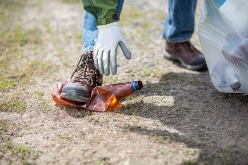 A person wearing green trousers, jeans, and brown boots is picking up a glass bottle from a dirt pathway outside, with their hand protected by a white glove. The bottle appears to be a repurposed brown glass with a blue cap, and it lies among loose soil and small patches of grass. In the background, a large white plastic rubbish bag is visible, likely filled with waste, and the scene takes place in an outdoor setting typical of rural or suburban areas, possibly in Mortlake or nearby in southwest London. The lighting is natural, indicating daytime, and the overall scene relates to rubbish collection or cleanup activities, highlighting the importance of safe waste handling supported by companies such as Office Clearance Mortlake.