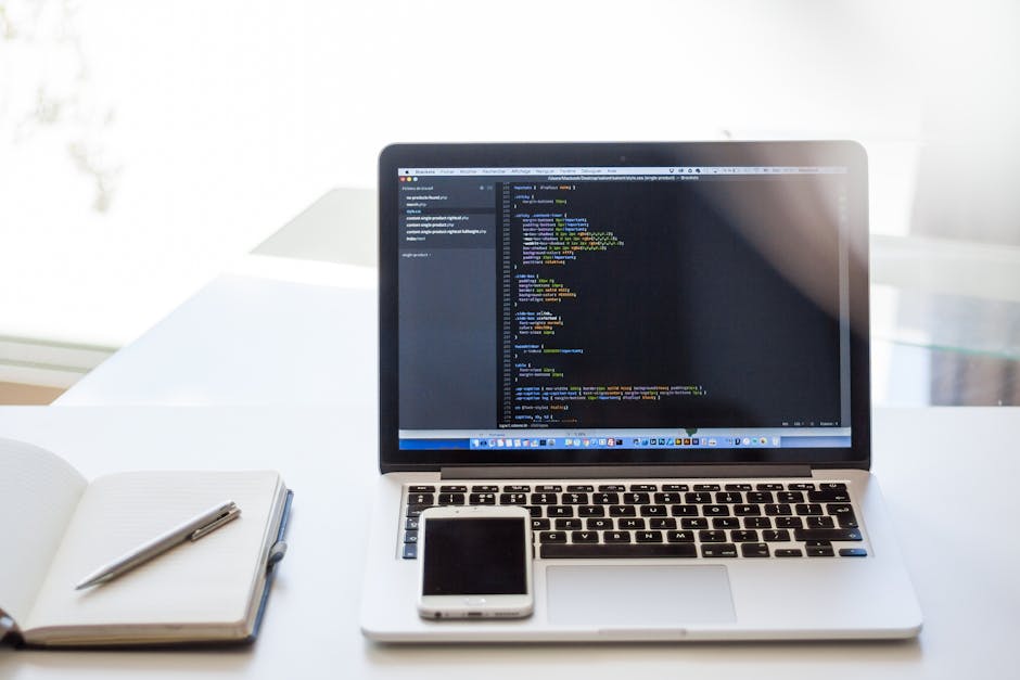 A silver MacBook laptop open on a white desk displaying lines of colorful computer code on a dark background, with a white smartphone placed in front of it. To the left, there is a white notebook with
