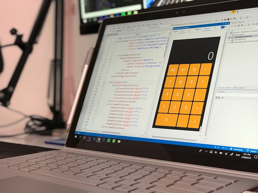 A silver MacBook laptop open on a white desk displaying lines of colorful computer code on a dark background, with a white smartphone placed in front of it. To the left, there is a white notebook with