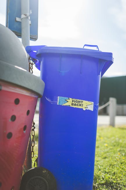 Five large wheeled rubbish bins with black plastic bodies and yellow lids are positioned in a row on a concrete surface against a plain, light-colored wall. Each bin has a white oval label with black 