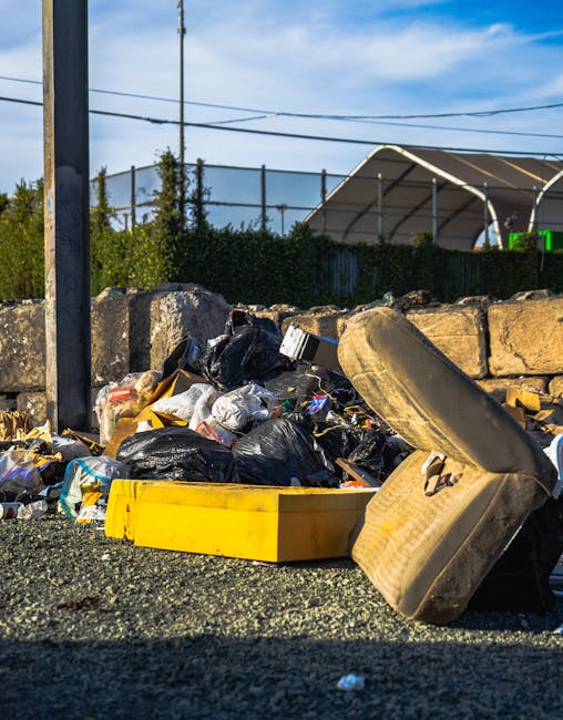 A collection of discarded waste materials piled on the ground in an outdoor area, including black plastic bags filled with rubbish, a yellow plastic container, and an old, dirty, beige upholstered arm