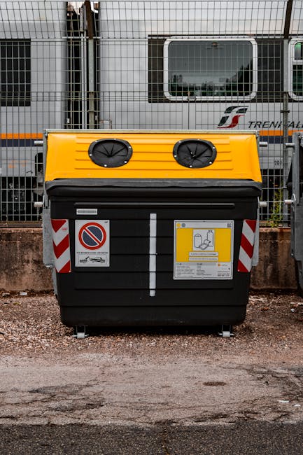 An overflowing collection of mixed waste and rubbish bags is piled around a grey and blue recycling bin labeled for paper and cardboard, situated on a paved area adjacent to a steel railing and a park